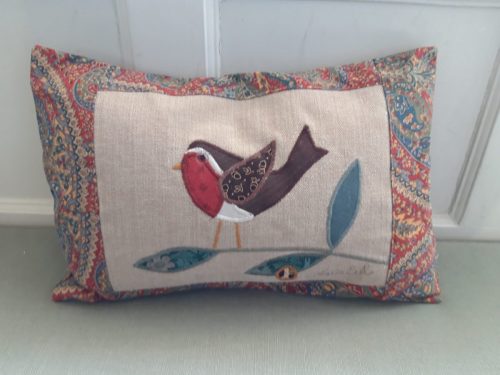 Robin cushion facing left in red and green paisley look border around a smooth beige ground with appliqué. The back flaps over, no zip. Size 48 x 29 cm (18” x 12”)