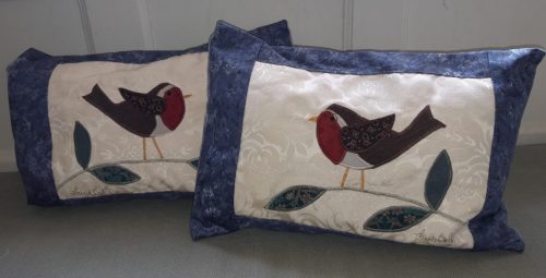 Pair of robin cushions, one facing right and one left. Mottled blue border around a pale cream slightly textured ground with appliqué. The backs are greyish blue with a swirly pattern and flap over, no zips. Size 48 x 29 cm (18” x 12”)