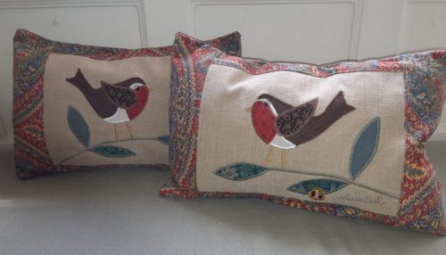 Pair of robin cushions, one facing right and one left. Red and green paisley look border around a pale cream slightly textured ground with appliqué. The backs flap over, no zips.
